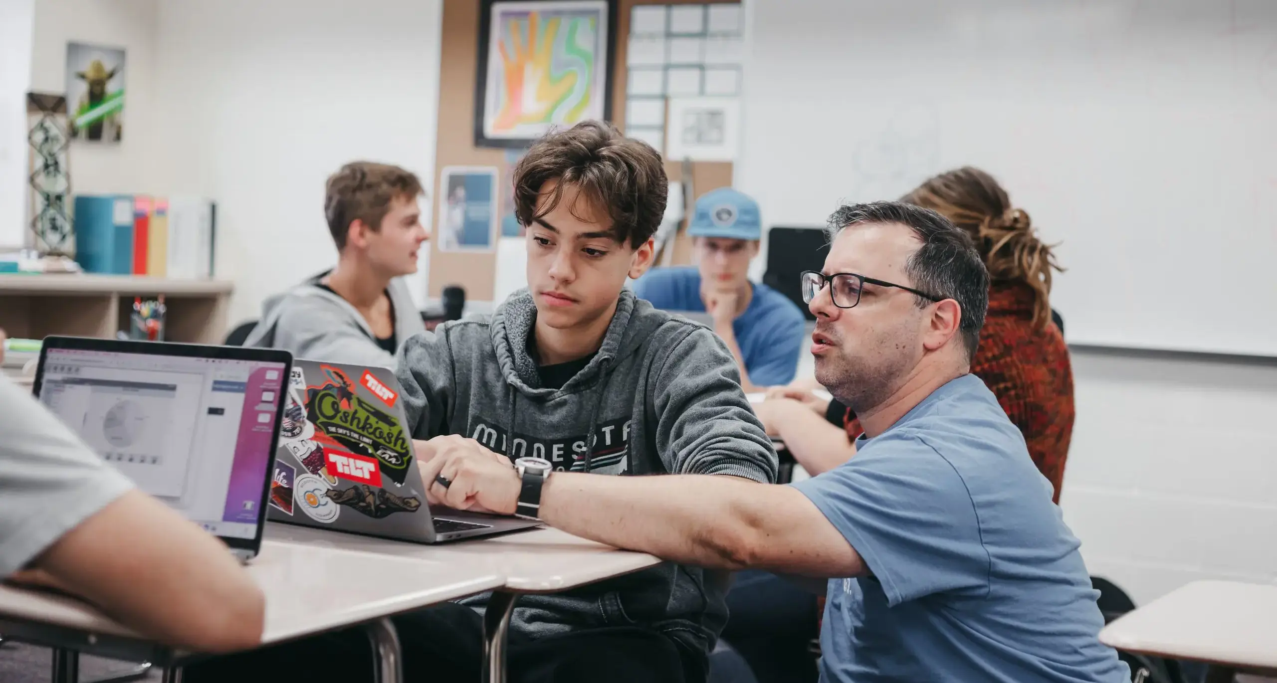 Teacher assisting a high school student with classwork on a laptop.