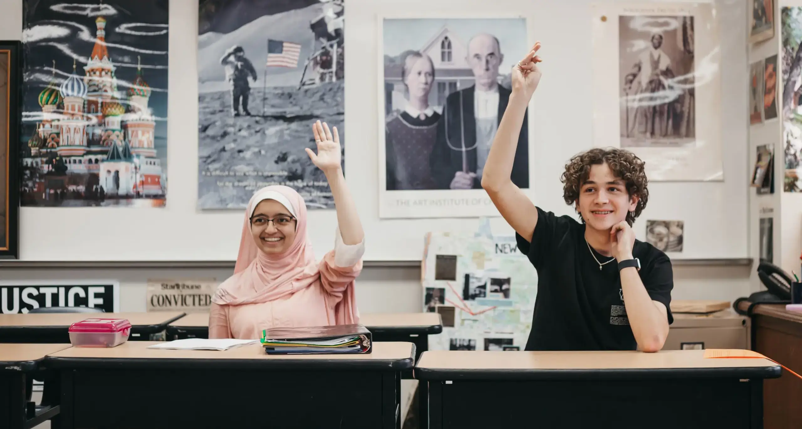 Two students seated in a classroom raise their hands during a lesson